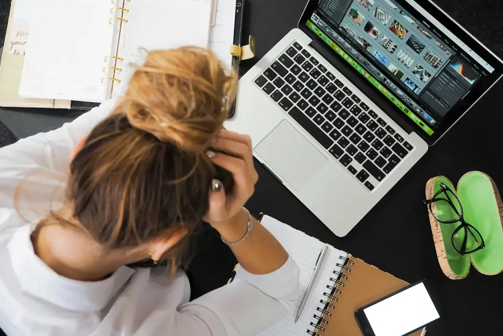 Woman sitting at a desk with her head in her hands, surrounded by a laptop, planner, notebook, and smartphone, highlighting work-related stress.